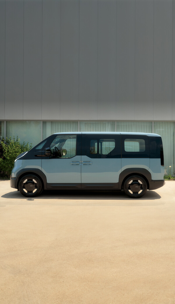 Vorderansicht des Kia PV5 Passenger, angeschlossen an eine Ladestation, mit einem Mann, der draußen neben Campingzubehör sitzt.